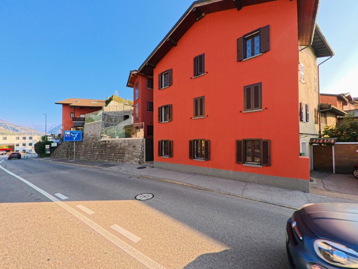 Entlang einer Strasse mit farbigen Wohnhaeusern zeigt sich ein gepflegtes Wohnquartier in Gravesano (6929, Kanton Tessin) bei klarem Himmel.