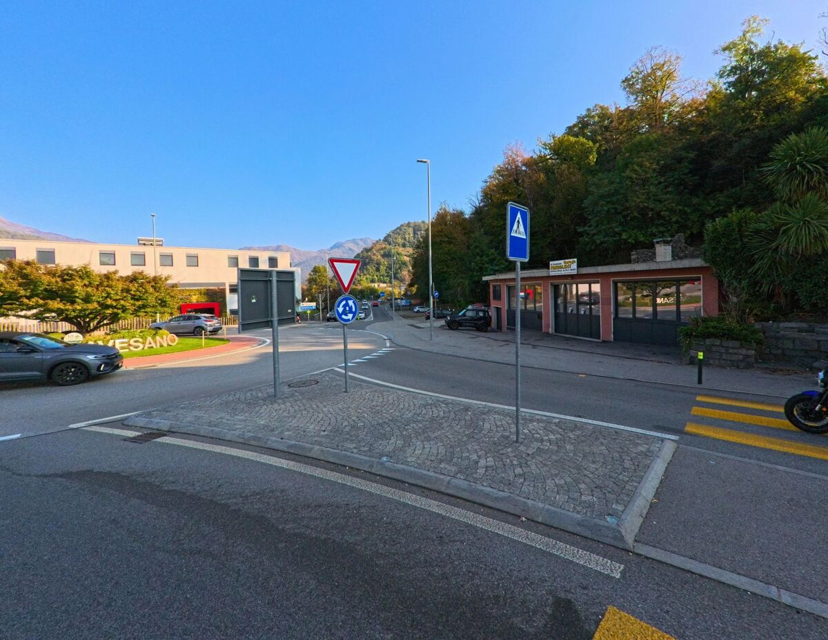 An einem Kreisverkehr mit Fussgaengerueberweg und Verkehrsschildern zeigt sich eine belebte Strassenszene in Gravesano (6929, Kanton Tessin) vor bewaldeten Haengen.