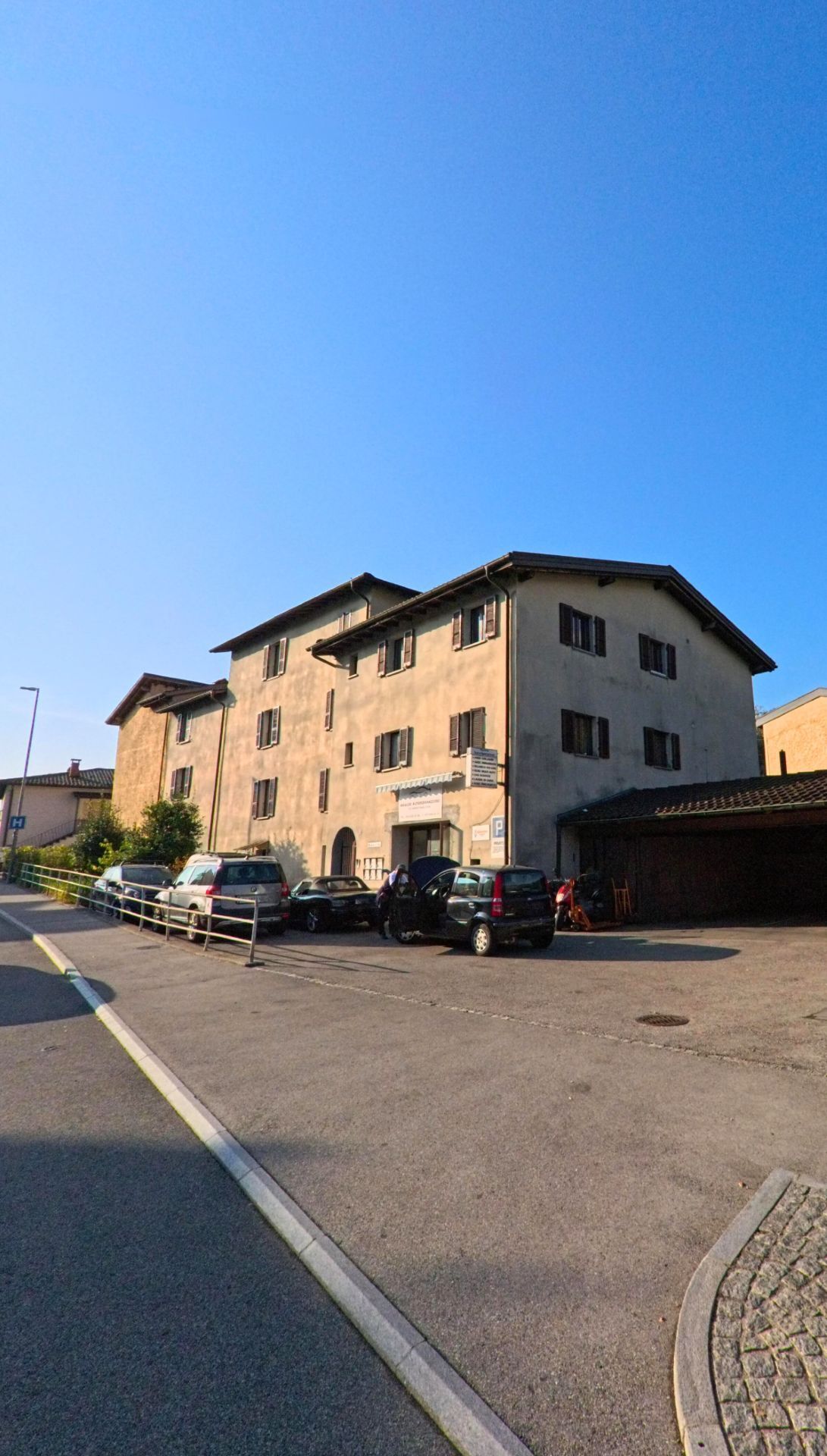 Ein mehrgeschossiges Gebaeude mit parkenden Autos praegt das Strassenbild in Gravesano (6929, Kanton Tessin) an einem sonnigen Tag.