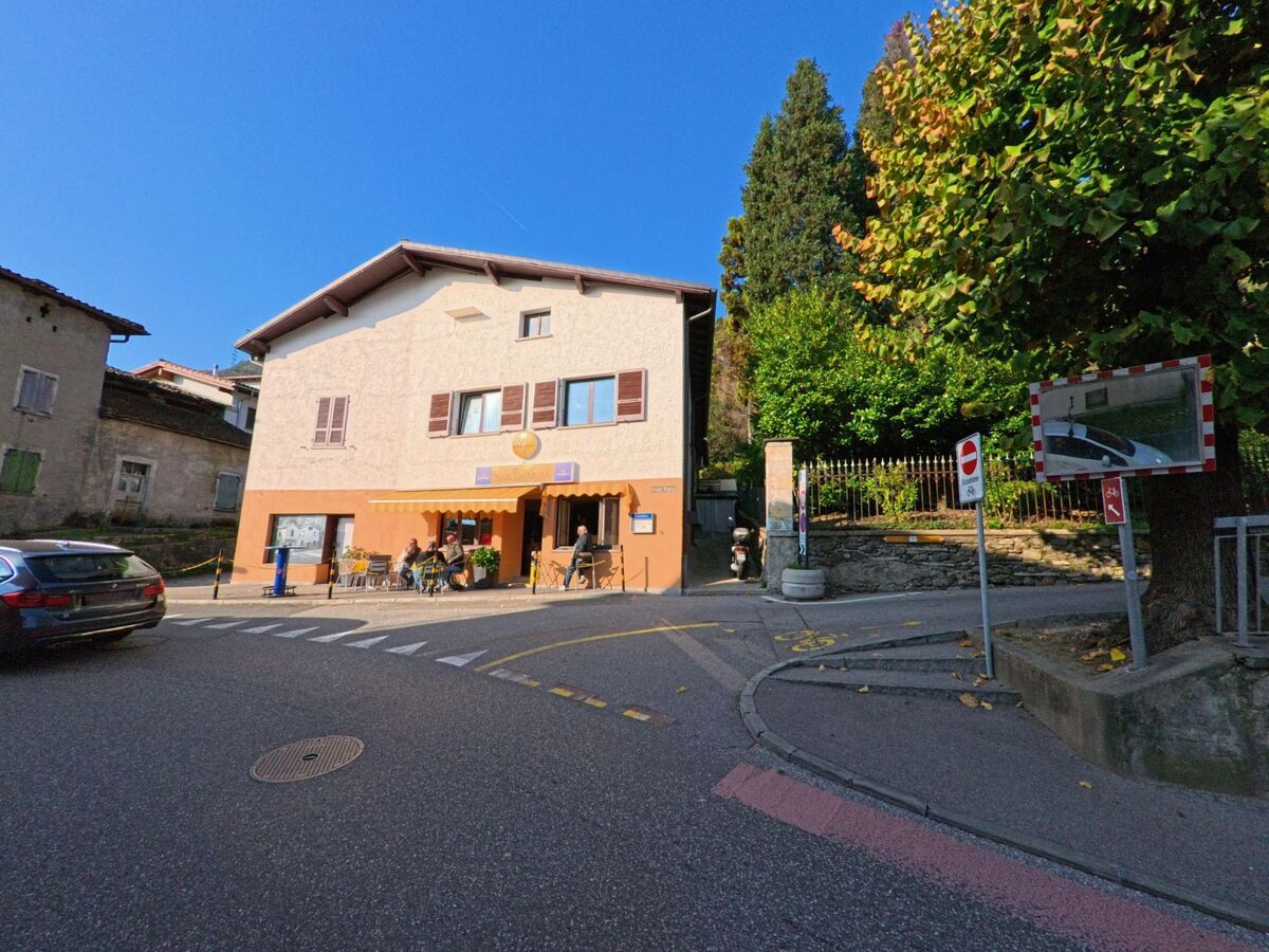 An einer Strassenecke mit Sitzplaetzen im Freien befindet sich ein kleines Restaurant in Gravesano (6929, Kanton Tessin), umgeben von Wohnhaeusern und Baeumen.