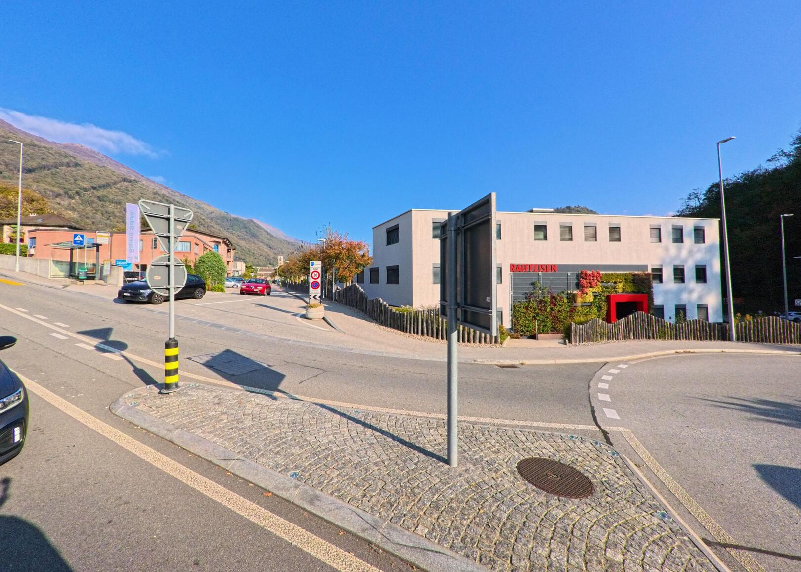 An einer Strassenkreuzung mit Verkehrsschildern steht ein modernes Bankgebaeude in Gravesano (6929, Kanton Tessin) vor bewaldeten Haengen.