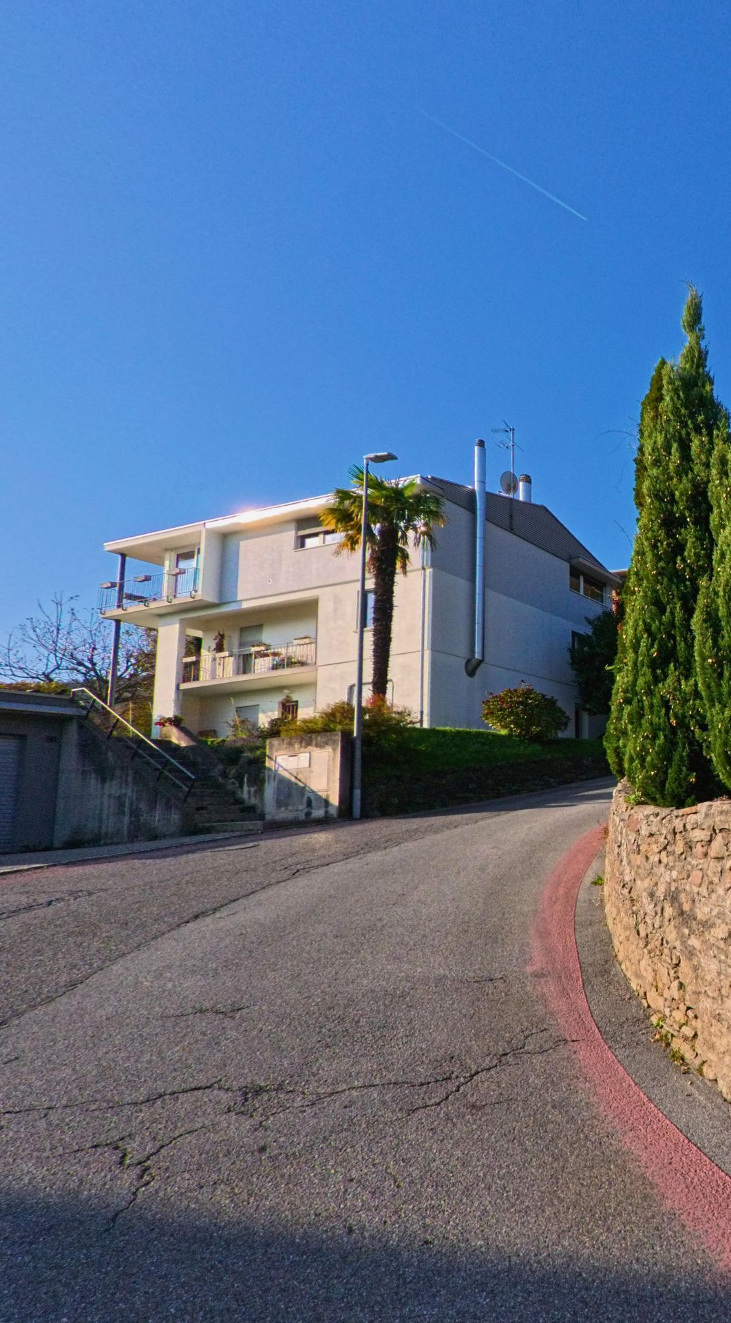 Ein modernes Wohnhaus steht an einer ansteigenden Strasse in Gravesano (6929, Kanton Tessin) mit Palmen und gepflegten Gruenflaechen unter blauem Himmel.