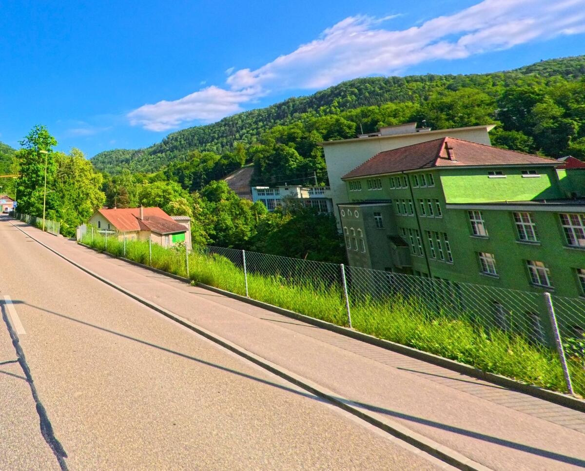 In Grellingen (4203, Kanton Baselland) führt eine Ausfallstrasse zwischen einem bewaldeten Hang und einem ehemaligen Industriebau in Richtung Tal.