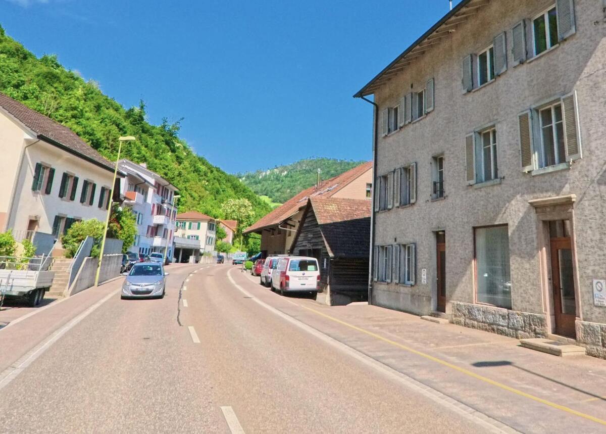 In Grellingen (4203, Kanton Baselland) verläuft eine Dorfstrasse mit dicht aneinandergereihten Wohnhäusern und parkenden Fahrzeugen entlang eines bewaldeten Hangs.