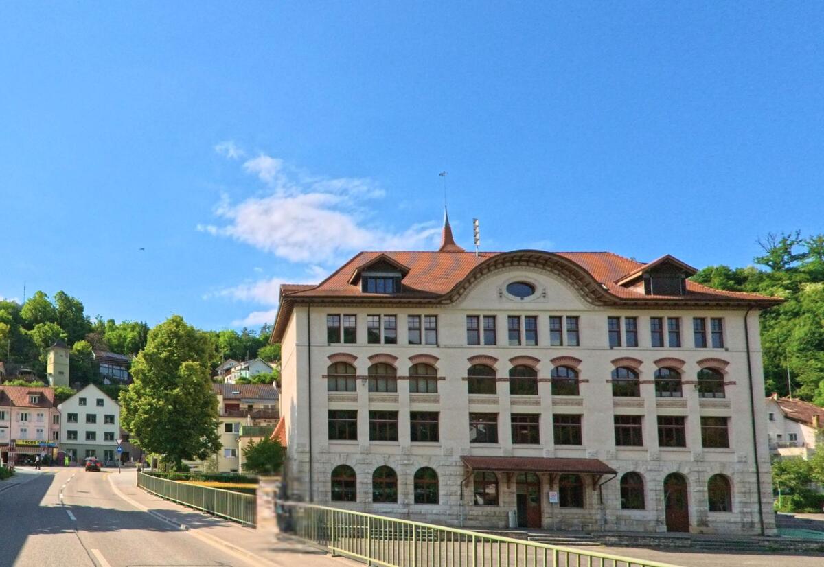 In Grellingen (4203, Kanton Baselland) steht ein markantes historisches Gebäude mit Rundbogenfenstern an einer Hauptstrasse, flankiert von weiteren Häusern und grünen Hügeln.