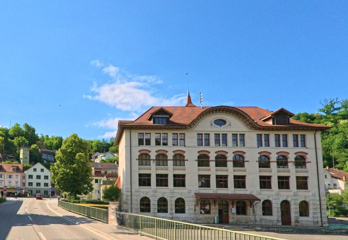 In Grellingen (4203, Kanton Baselland) steht ein markantes historisches Gebäude mit Rundbogenfenstern an einer Hauptstrasse, flankiert von weiteren Häusern und grünen Hügeln.