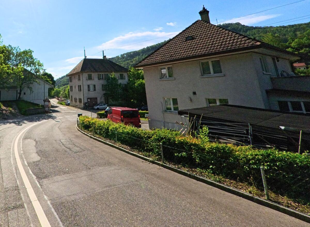 In Grellingen (4203, Kanton Baselland) verläuft eine leicht abschüssige Strasse zwischen Wohnhäusern und einer Baustelle mit Baukran am Waldrand.