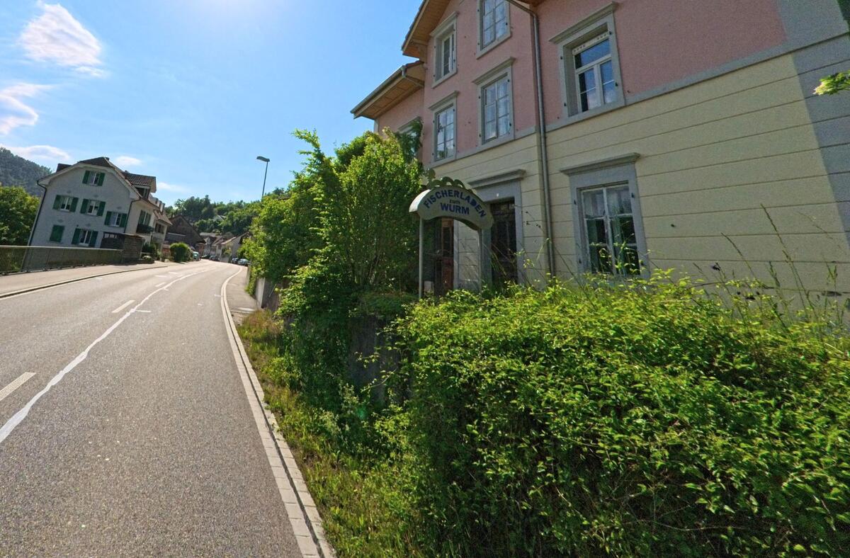 In Grellingen (4203, Kanton Baselland) führt eine sonnige Strasse an einem rosa gestrichenen Gebäude mit der Aufschrift „Bohrerlager Wurm“ vorbei und überquert dabei eine Brücke.