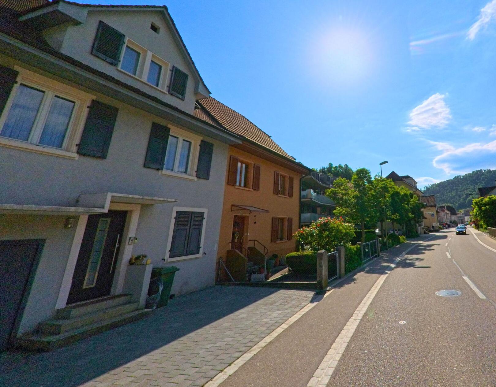In Grellingen (4203, Kanton Baselland) verläuft eine sonnige Wohnstrasse mit Reihenhäusern und Gehweg entlang einer leichten Kurve in Richtung Ortszentrum.