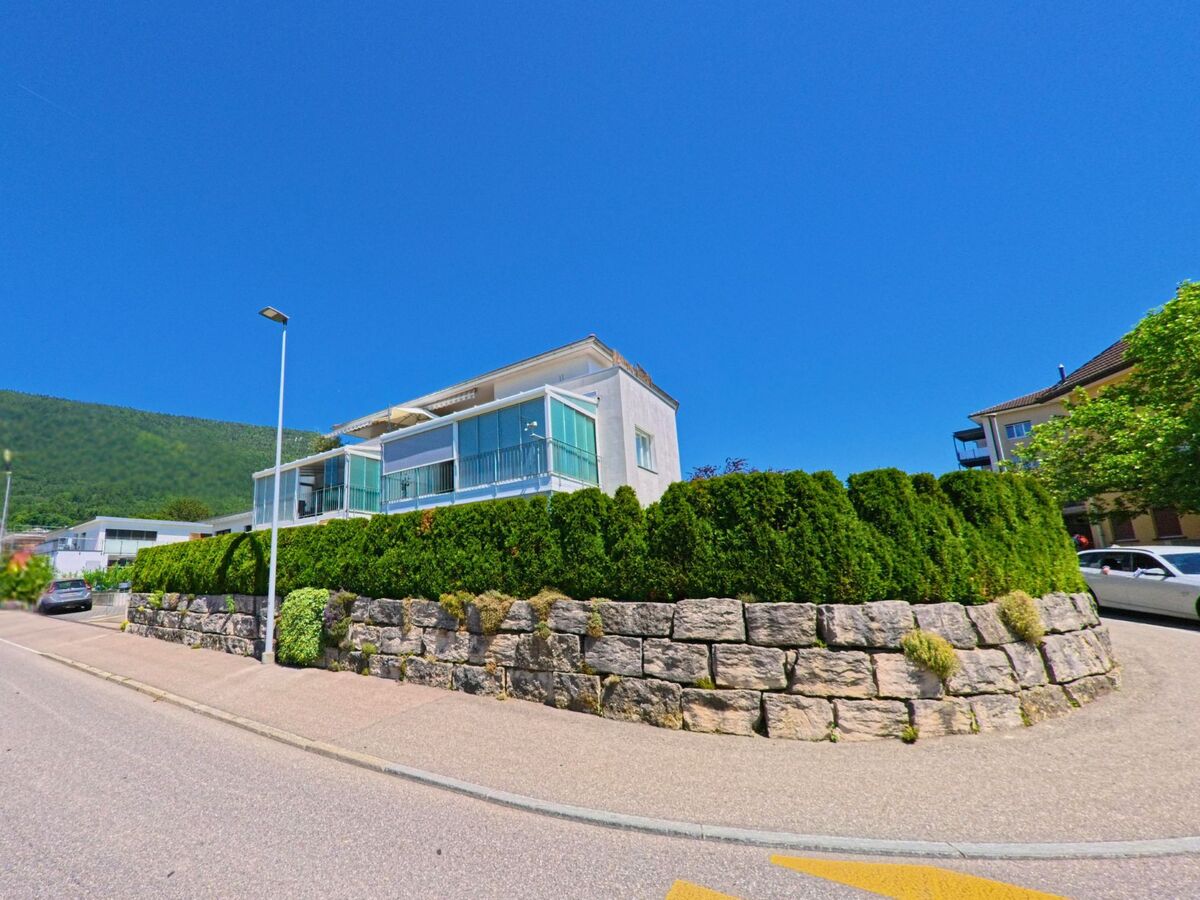 In Grenchen (2540, Kanton Solothurn) steht ein modernes Wohnhaus mit verglasten Balkonen oberhalb einer Natursteinmauer und gepflegter Hecke am Strassenrand.