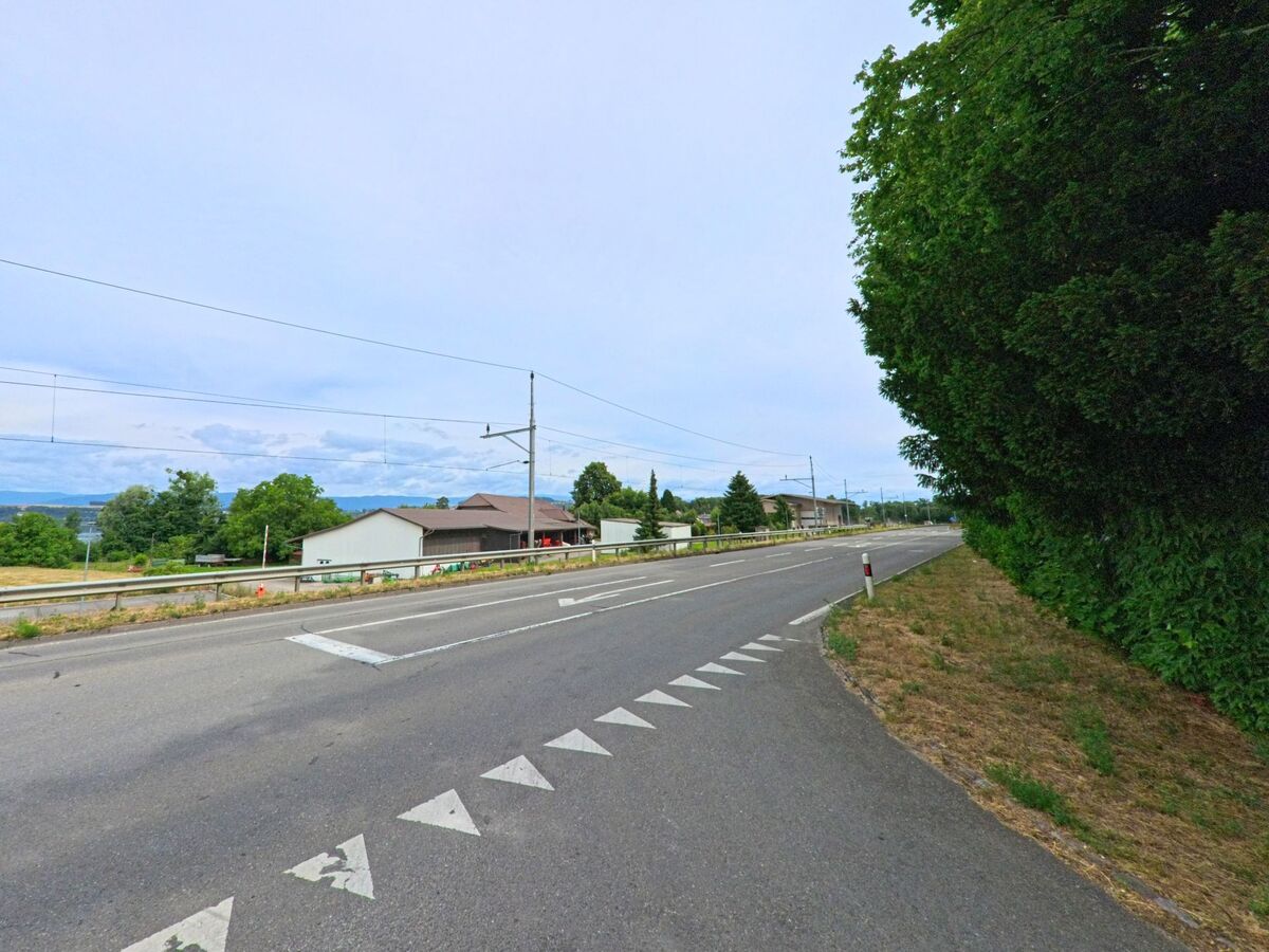 Entlang einer breiten Durchgangsstrasse in Greng (3280, Kanton Freiburg) verläuft eine Bahnlinie mit Oberleitungen, während auf der linken Seite landwirtschaftliche Gebäude zu sehen sind.