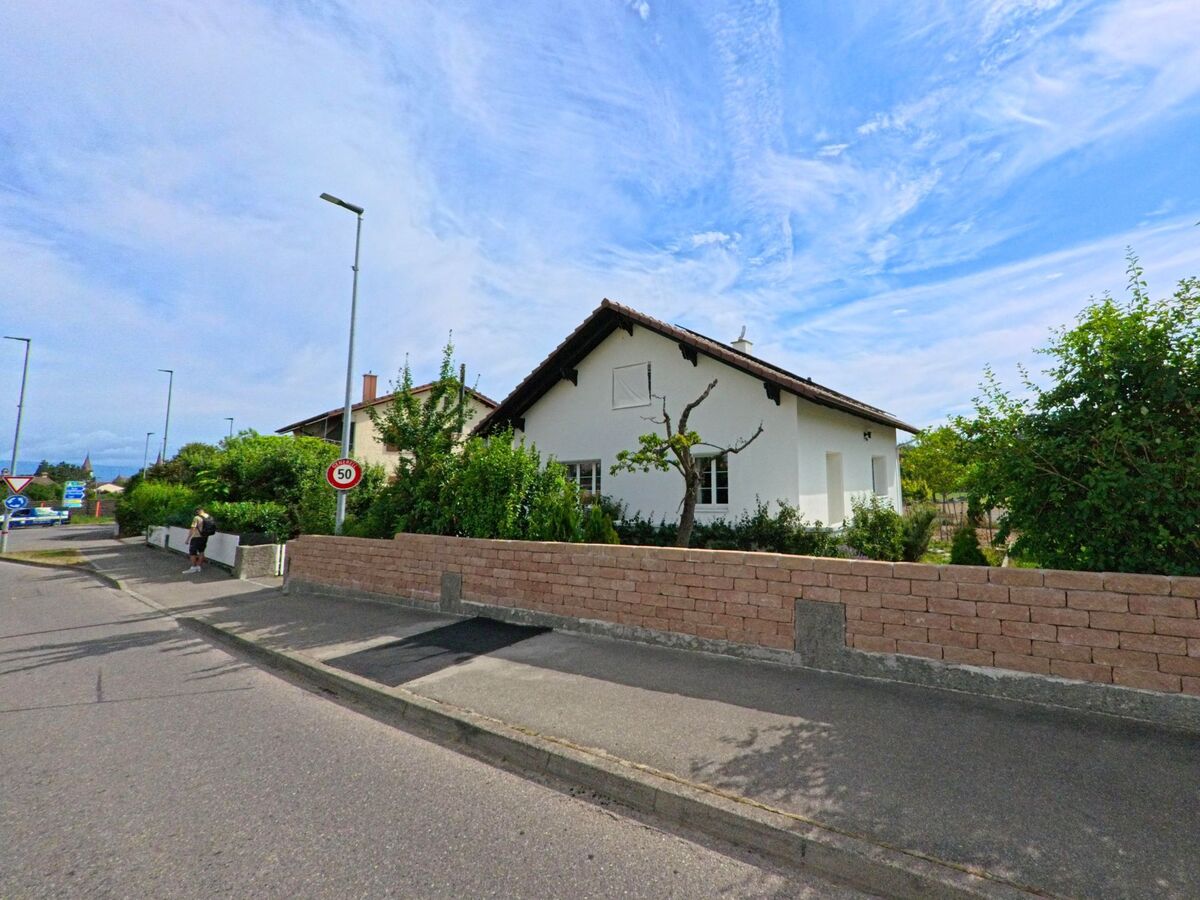 In Greng (3280, Kanton Freiburg) liegt ein weiss gestrichenes Einfamilienhaus mit gepflegtem Garten hinter einer niedrigen Mauer an einer verkehrsberuhigten Strasse.