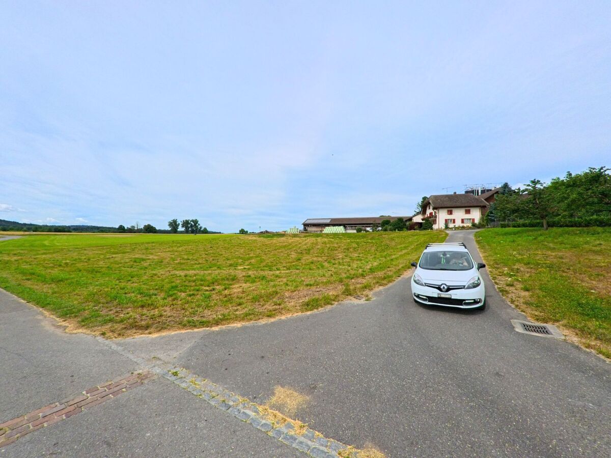 Ein schmaler Weg führt in Greng (3280, Kanton Freiburg) über offene Felder zu einem Bauernhaus mit roten Fensterläden, während ein Auto in Richtung Kamera fährt.
