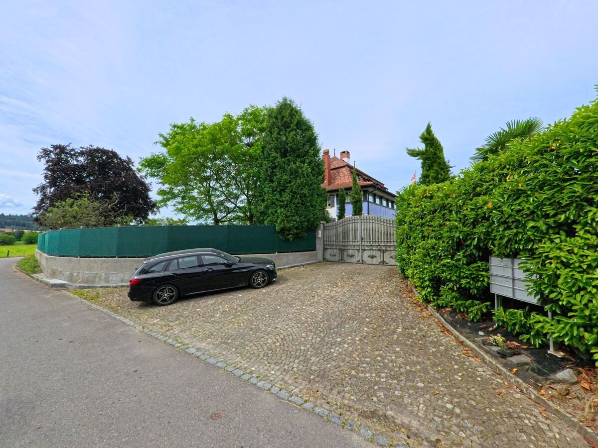 Am Ende einer gepflasterten Zufahrt in Greng (3280, Kanton Freiburg) steht ein herrschaftliches Wohnhaus mit rotem Dach, umgeben von hohen Hecken und Bäumen.