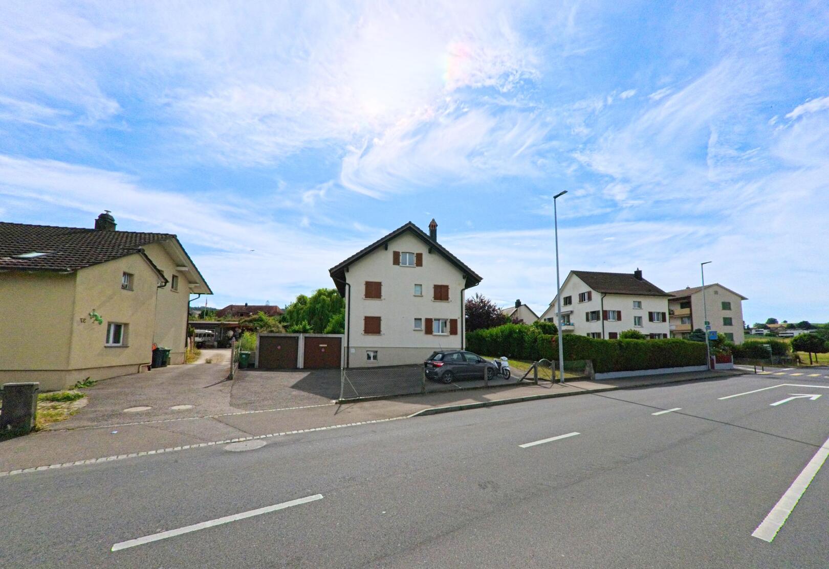 In Greng (3280, Kanton Freiburg) stehen entlang einer breiten Strasse mehrere Mehrfamilienhäuser mit gepflegten Vorplätzen und Garagenanlagen.