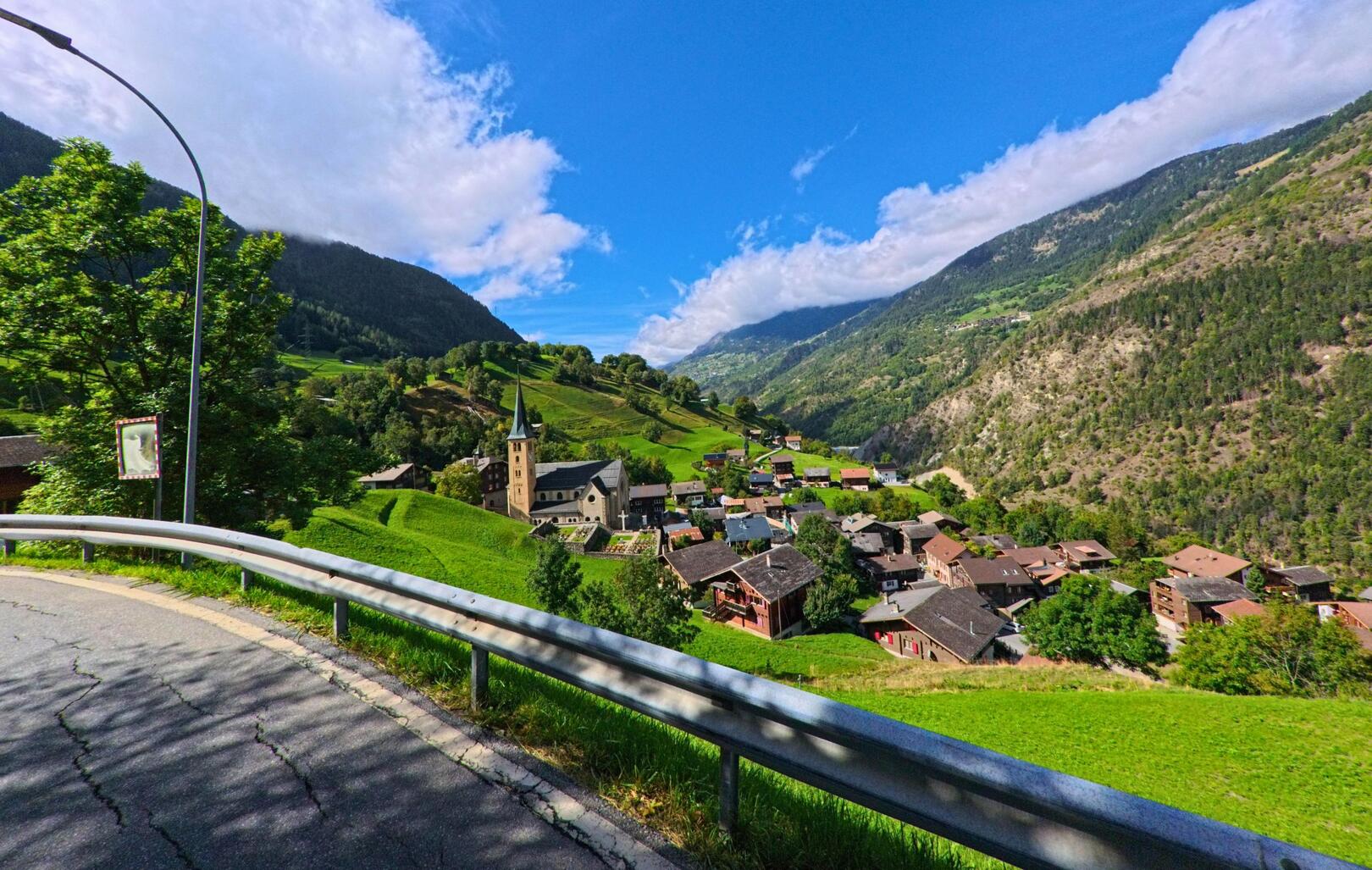 Vom Strassenrand aus bietet sich ein weiter Blick auf Grengiols (3993, Kanton Wallis) mit der Kirche im Zentrum und den umgebenden Bergen.