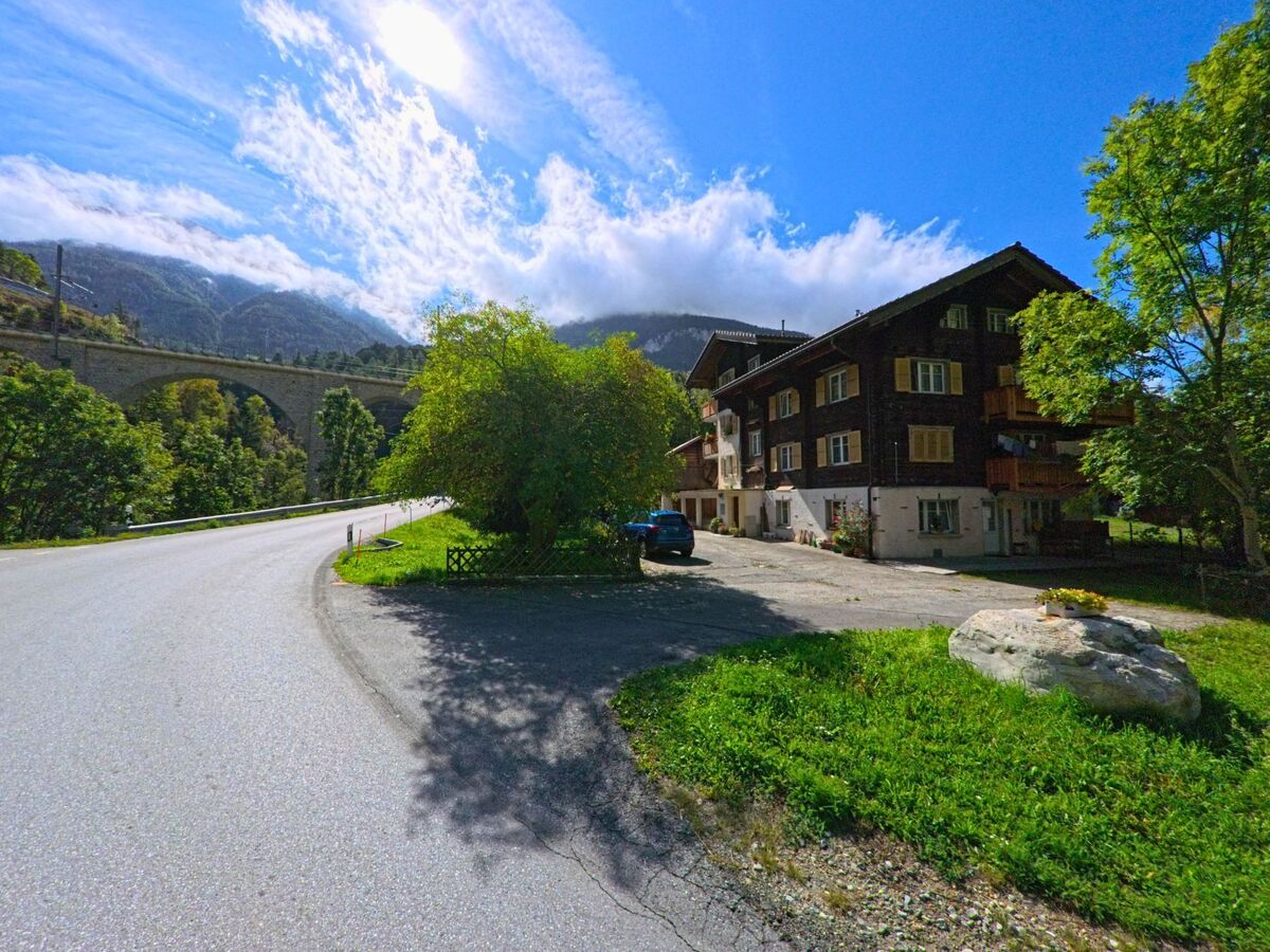 Am Ortsrand von Grengiols (3993, Kanton Wallis) steht ein traditionelles Wohnhaus nahe der Strasse, während im Hintergrund ein steinernes Eisenbahnviadukt sichtbar ist.