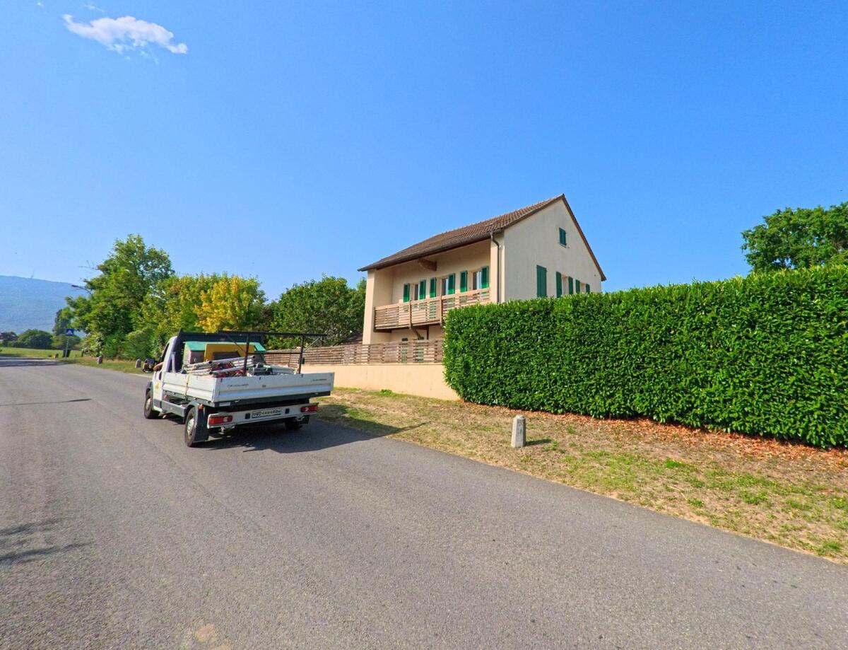 Ein Wohnhaus mit gruenen Fensterlaeden in Grens (1274, Kanton Waadt) steht hinter einer hohen Hecke an einer ruhigen Strasse.