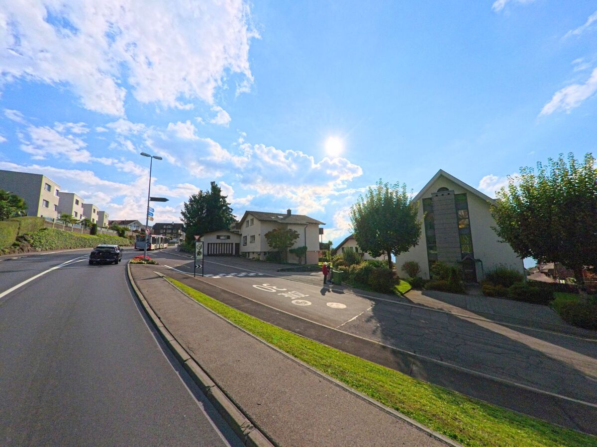 In Greppen (6404, Kanton Luzern) fuehrt eine sanft ansteigende Strasse an modernen und traditionellen Wohnhaeusern vorbei unter einem klaren Himmel.