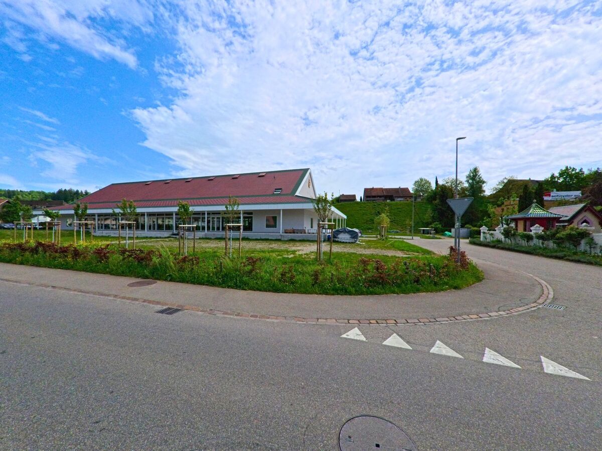 In Gretzenbach (5014, Kanton Solothurn) liegt ein modernes, flach gebautes Gebäude mit rotem Dach an einer Verkehrsinsel mit jungen Bäumen und Grünfläche.