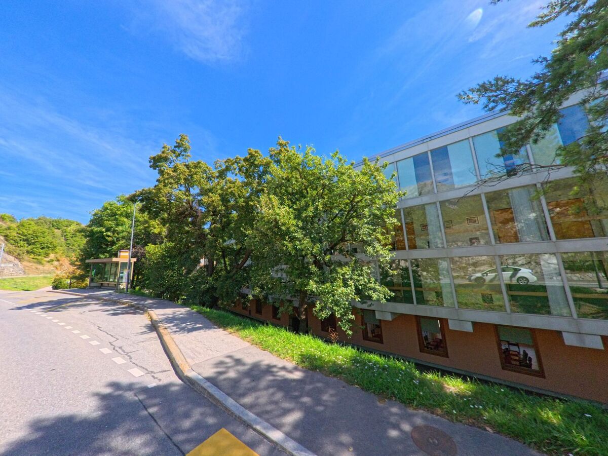 Ein modernes Gebaeude mit grossen Glasfassaden steht in Grimisuat (1971, Kanton Wallis) neben einer Strasse mit Bushaltestelle.