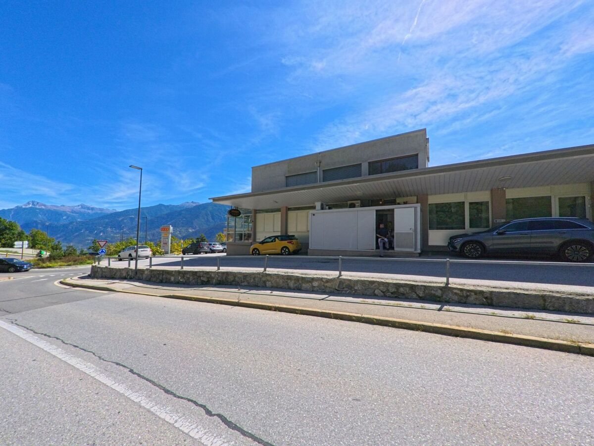 Ein flaches, modernes Gebaeude mit Parkplaetzen davor steht an einer Strasse in Grimisuat (1971, Kanton Wallis) mit Blick auf die Berge.