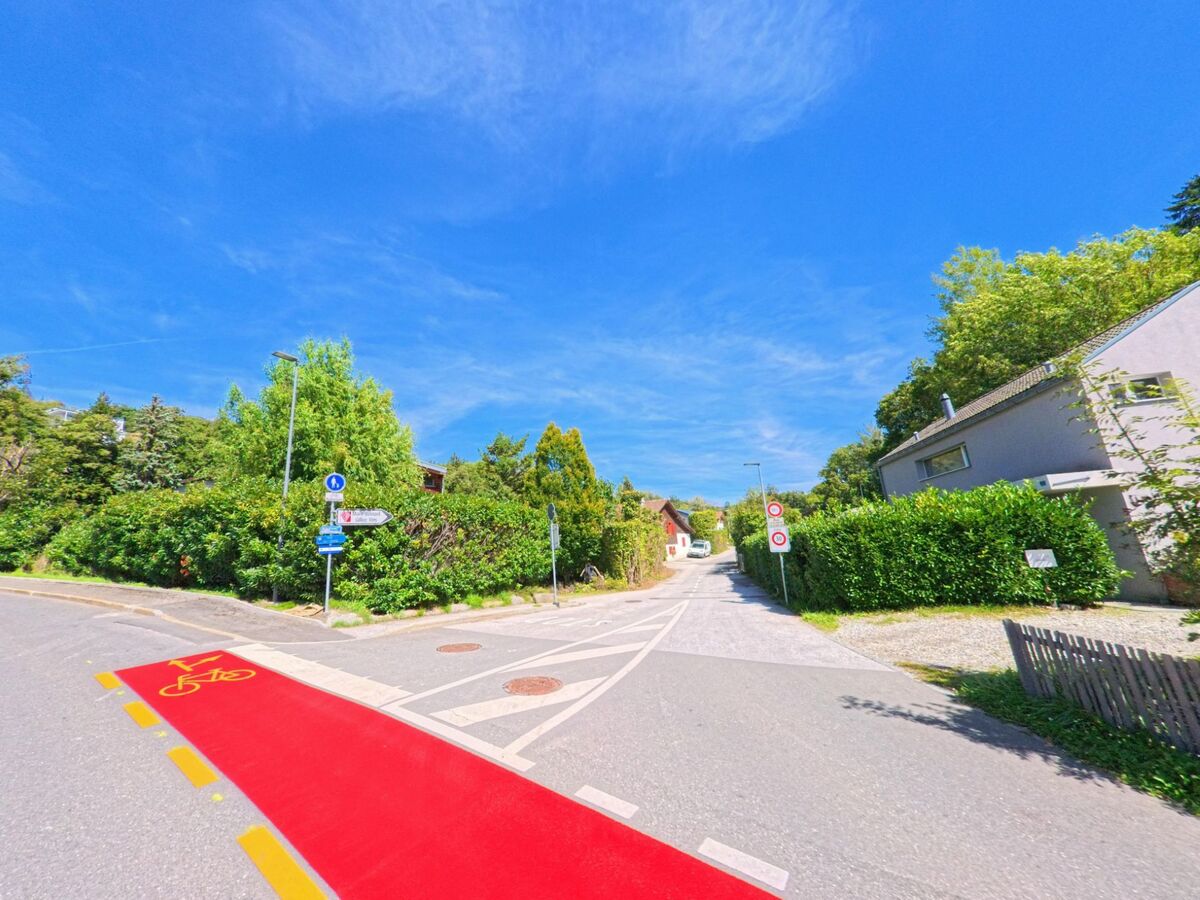 Ein rot markierter Radstreifen fuehrt in Grimisuat (1971, Kanton Wallis) ueber die Strasse, daneben beginnt ein schmaler Weg mit Wohnhaeusern.