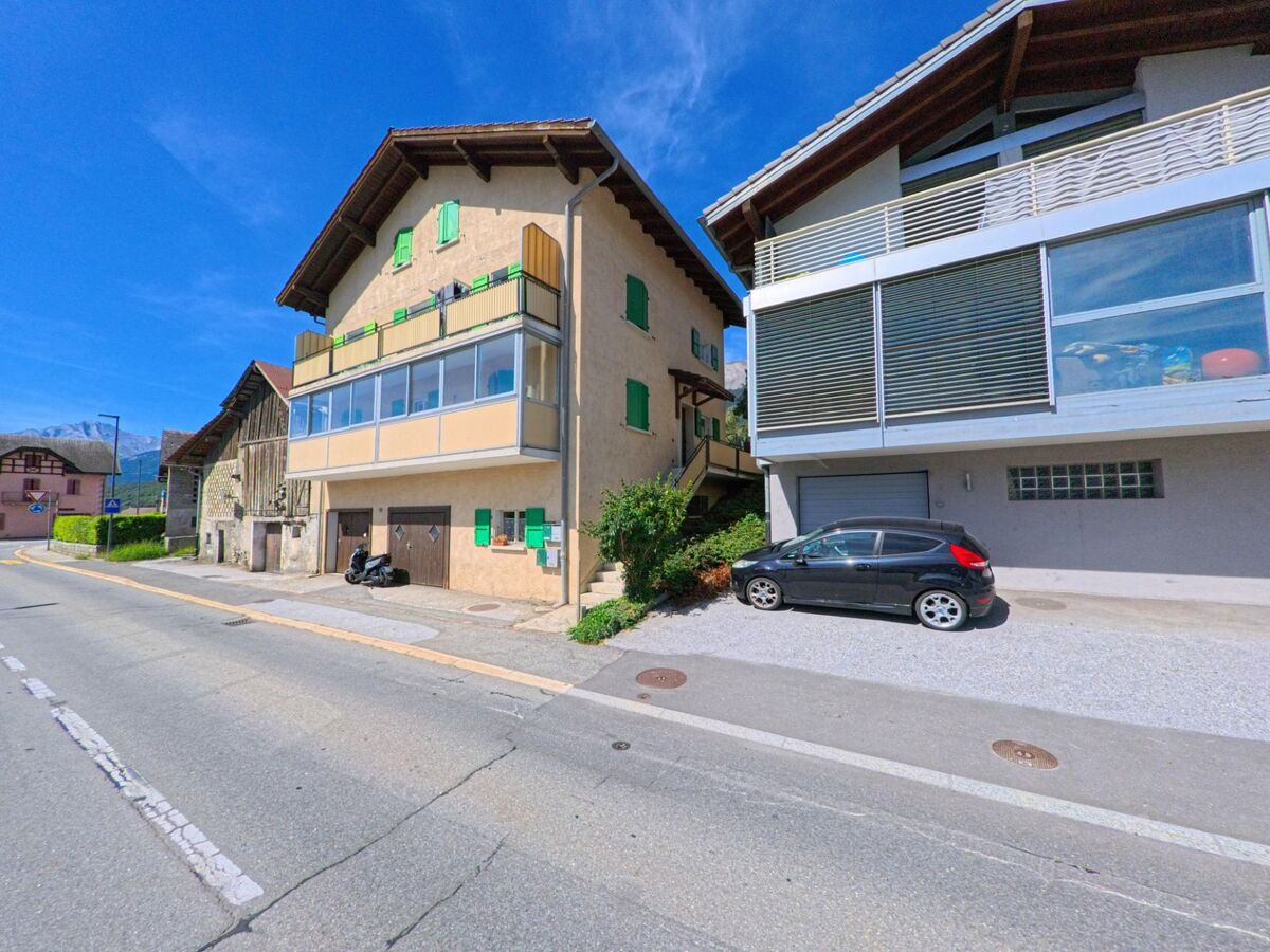 Ein Wohnhaus mit gruenen Fensterlaeden und verglastem Balkon steht an einer Strasse in Grimisuat (1971, Kanton Wallis).