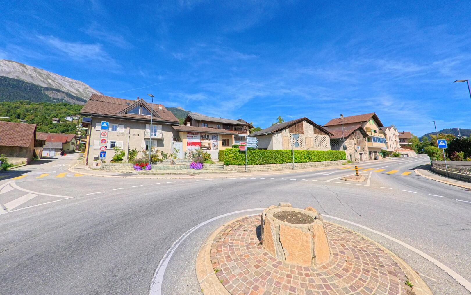 Eine Strassenkreuzung mit kleiner Verkehrsinsel liegt in Grimisuat (1971, Kanton Wallis) umgeben von Wohnhaeusern und Blick auf den Berghang.