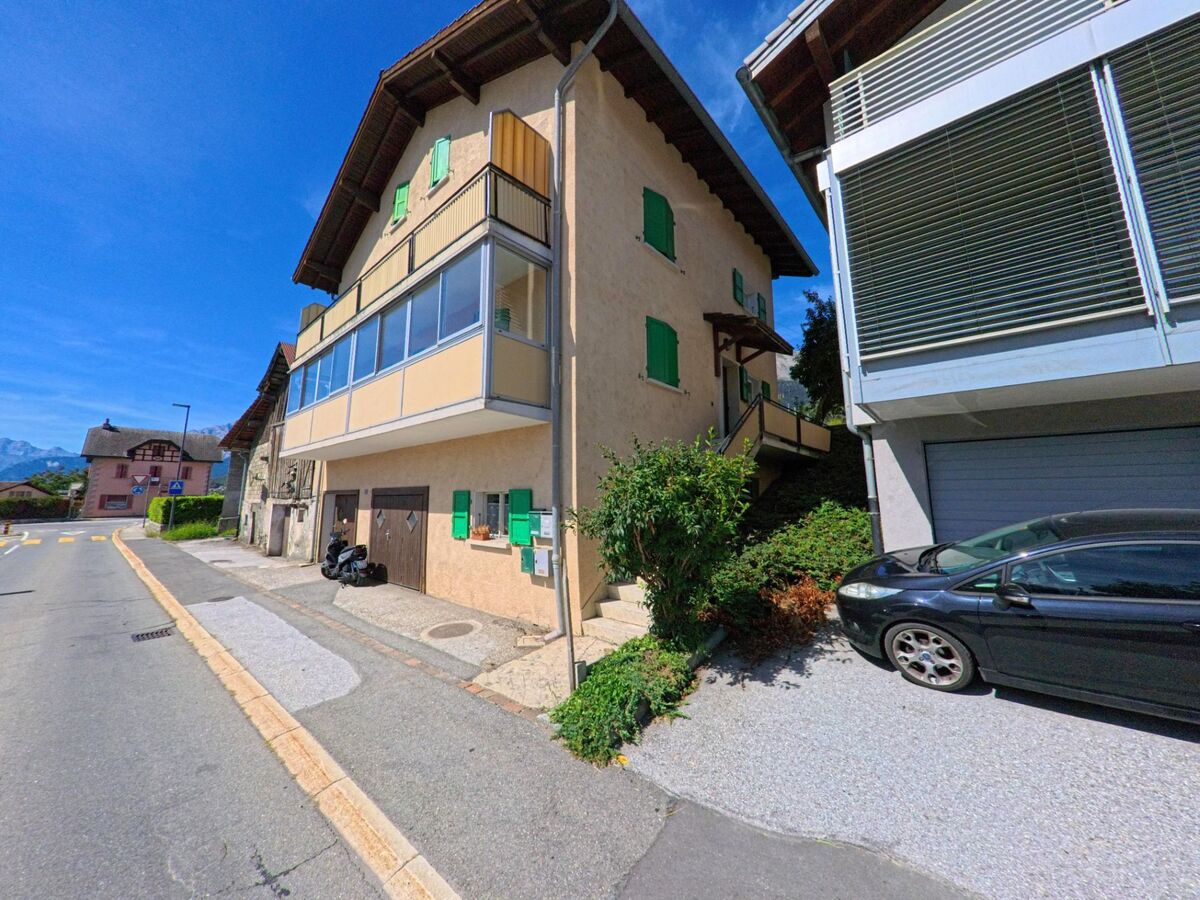 An einer Strasse in Grimisuat (1971, Kanton Wallis) stehen traditionelle und moderne Wohnhaeuser nebeneinander, mit Garagen und Balkonen.