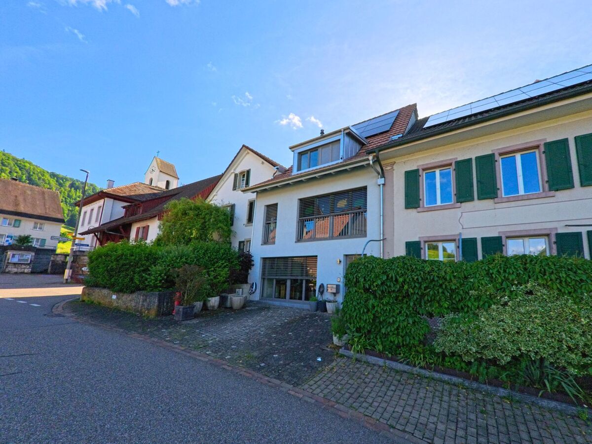 In Grindel (4247, Kanton Solothurn) stehen historische Wohnhäuser mit grünen Fensterläden und moderner Solaranlage nahe der Dorfkirche an einer Kurve.