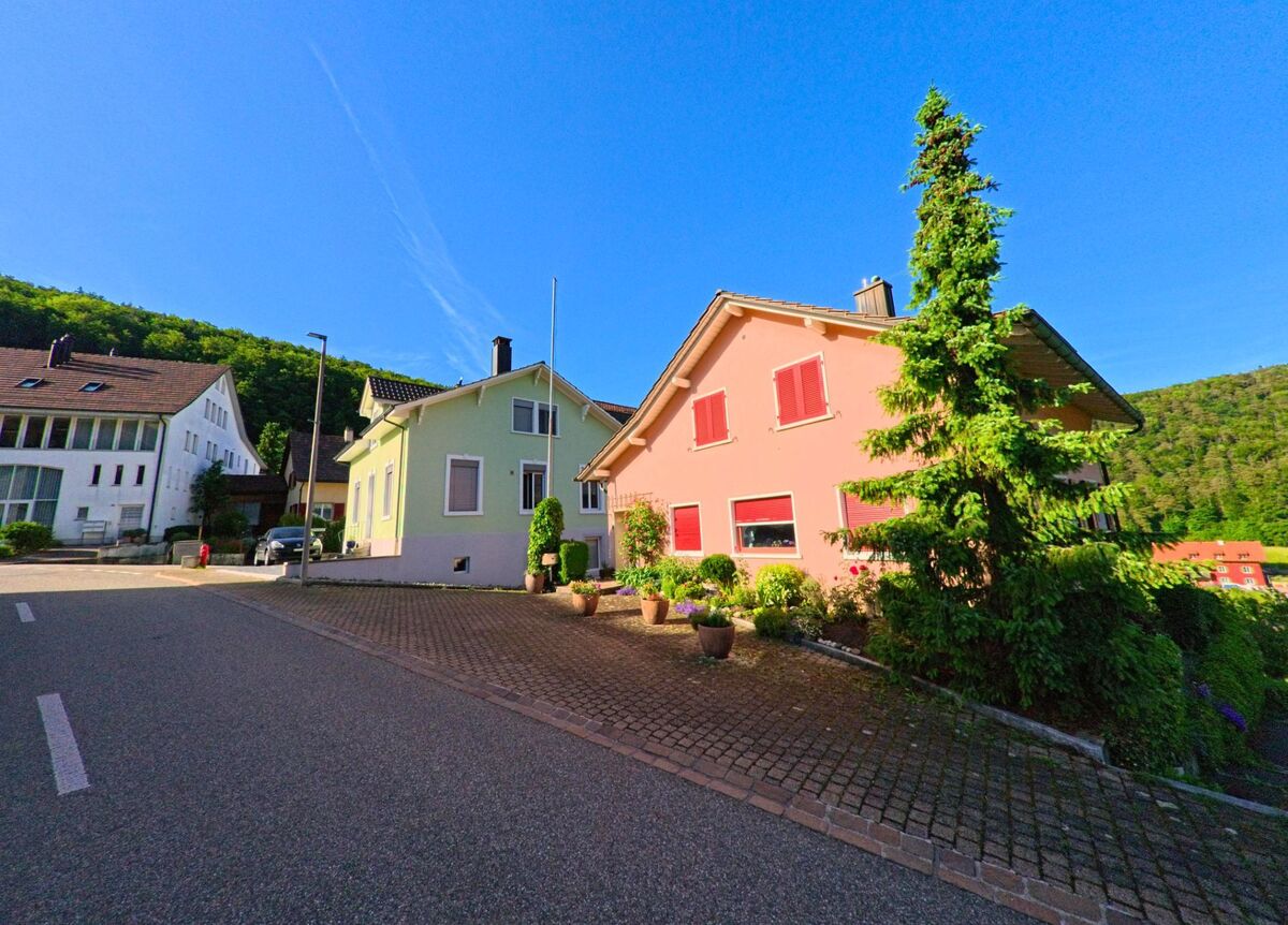 In Grindel (4247, Kanton Solothurn) stehen farbige Einfamilienhäuser mit gepflegten Vorgärten an einer sonnigen Strasse am Waldrand.