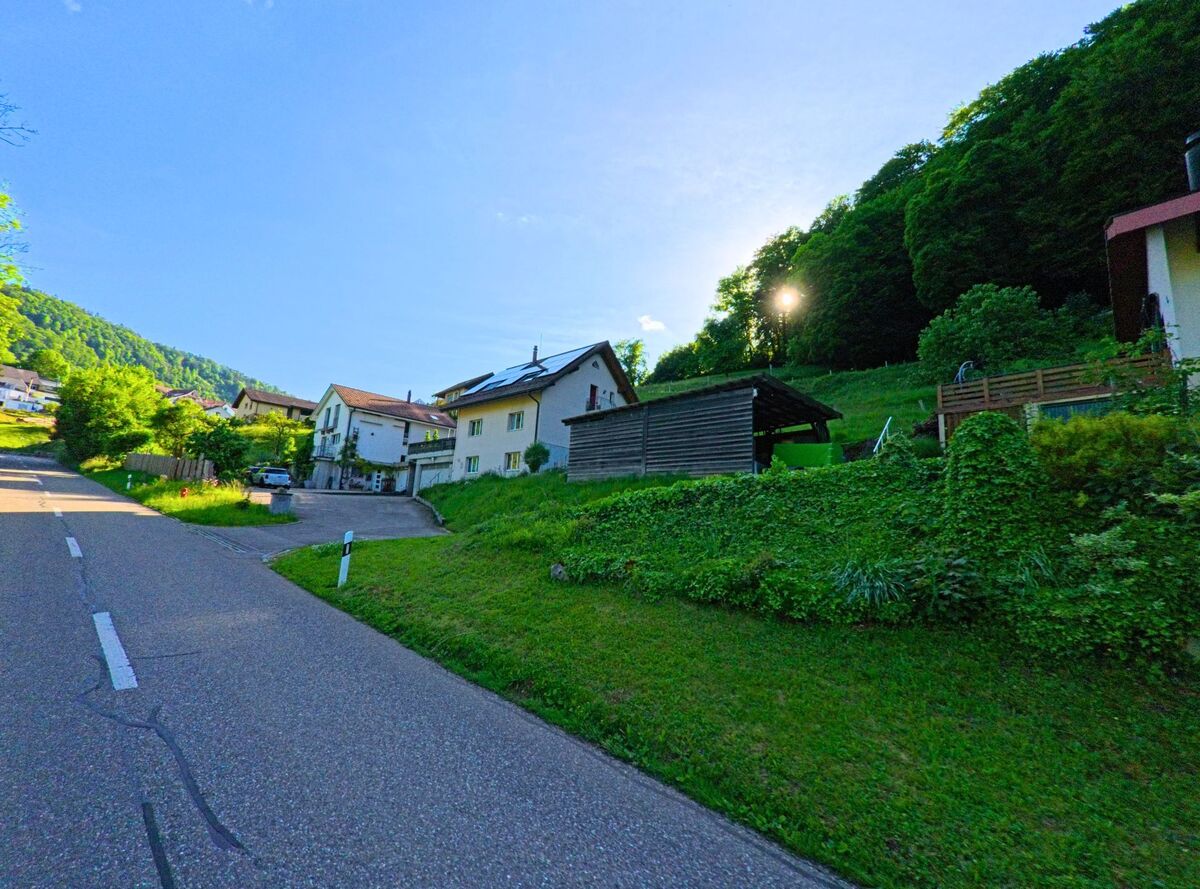 Entlang einer ruhigen Strasse in Grindel (4247, Kanton Solothurn) reihen sich Hanghäuser mit Gärten an einem bewaldeten Abhang.