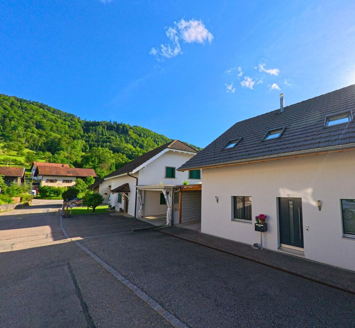 Am Fuss bewaldeter Hügel stehen in Grindel (4247, Kanton Solothurn) moderne Einfamilienhäuser entlang einer gepflegten Quartierstrasse im Sonnenlicht.