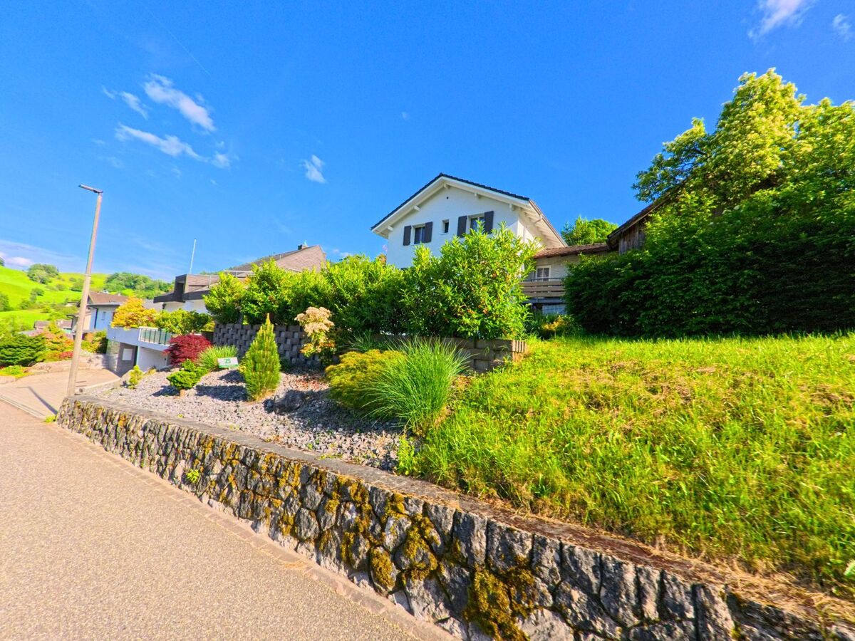  In Grindel (4247, Kanton Solothurn) säumen gepflegte Vorgärten und terrassierte Steinmauern eine ruhige Wohnstrasse mit Blick auf grüne Hügellandschaft.