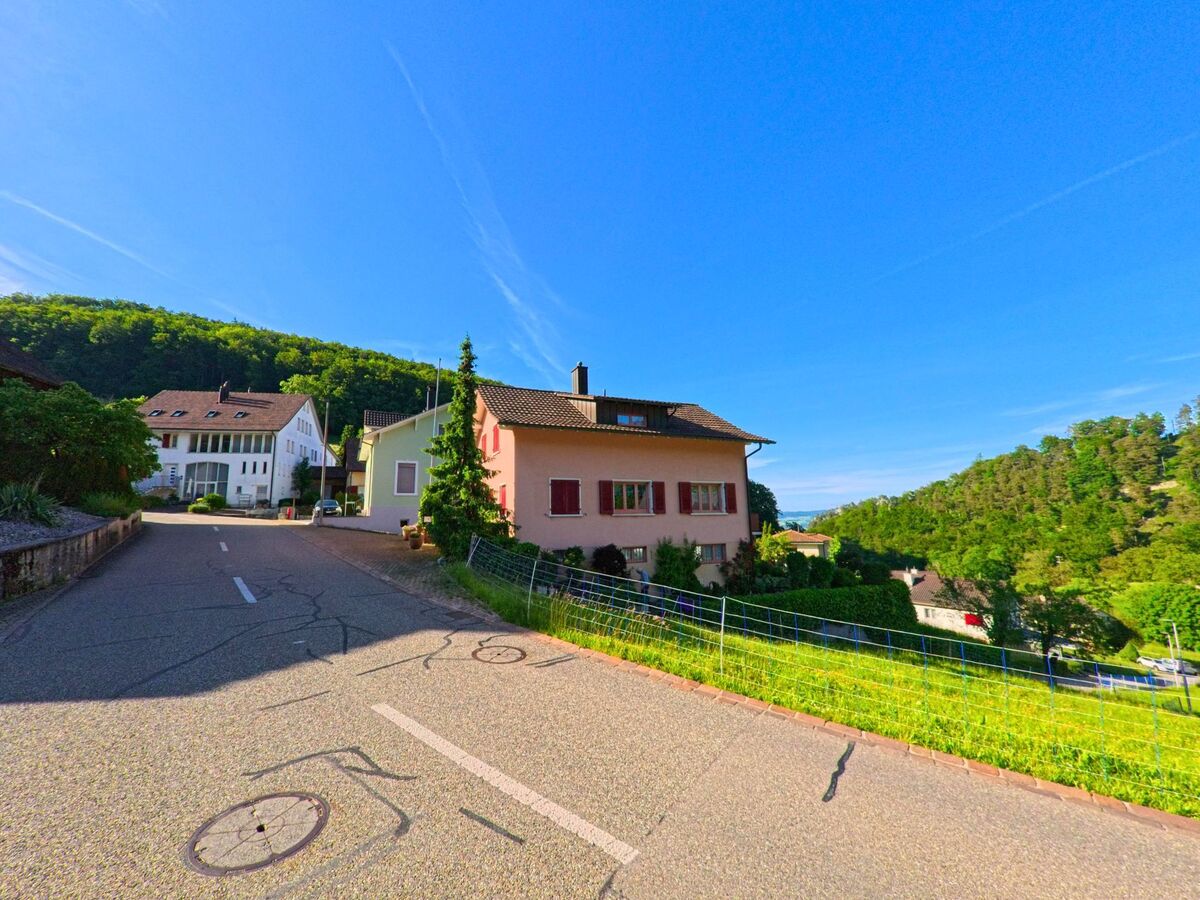 Eine ansteigende Strasse in Grindel (4247, Kanton Solothurn) führt an Wohnhäusern mit roten Fensterläden vorbei, flankiert von Wiesen und bewaldeten Hängen.