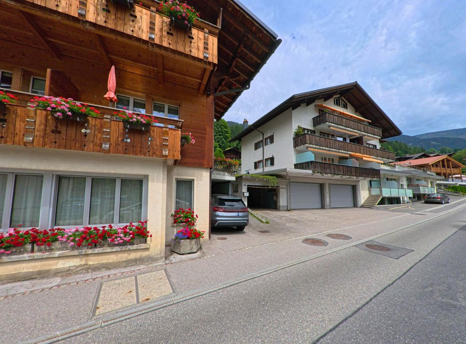 In Grindelwald (3818, Kanton Bern) reihen sich entlang einer leicht ansteigenden Strasse traditionelle und moderne Wohnhaeuser mit holzverkleideten Balkonen und farbenfrohem Blumenschmuck.