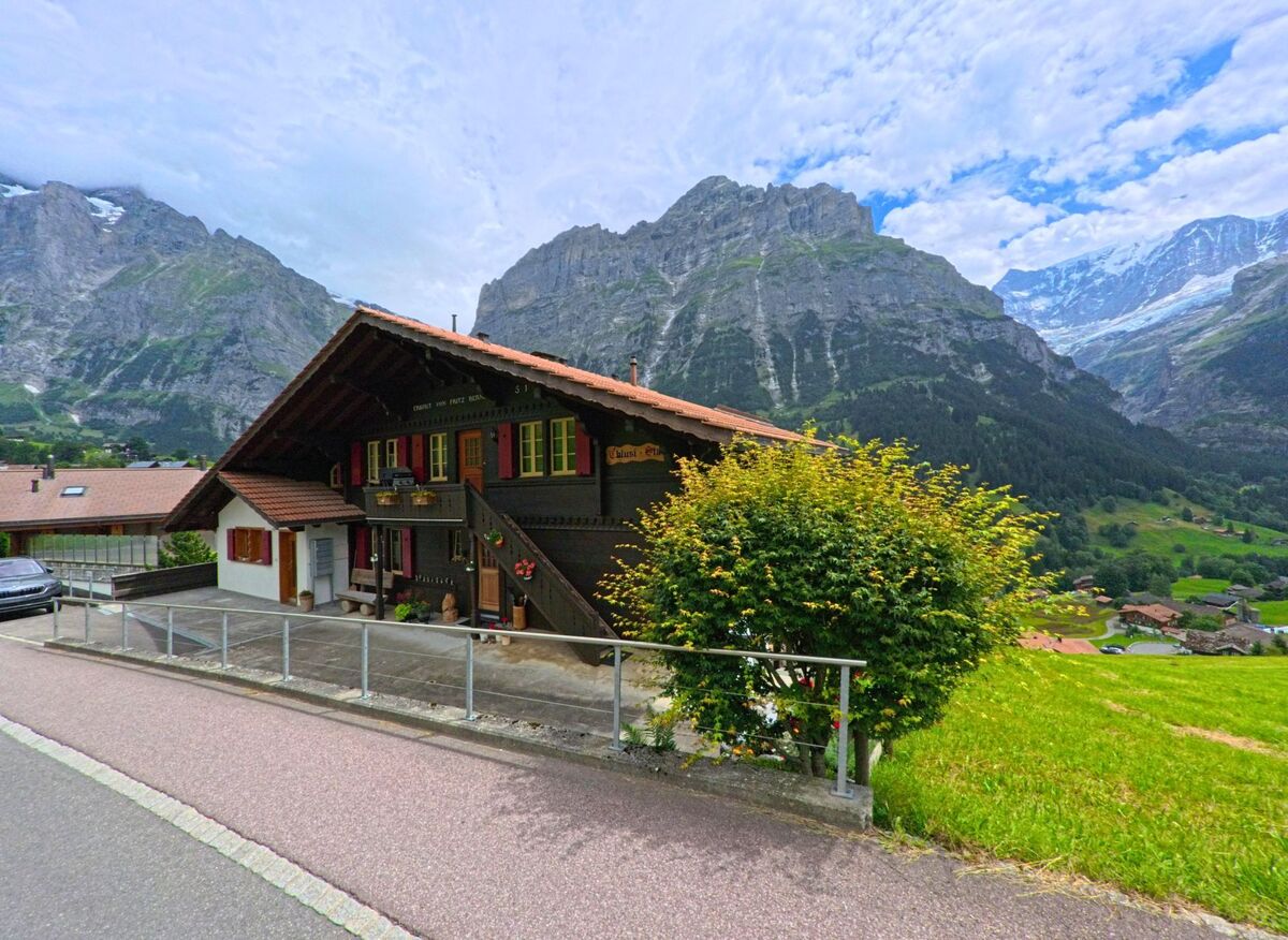 Vor der imposanten Bergkulisse von Grindelwald (3818, Kanton Bern) steht ein traditionelles Holzhaus mit farbigen Fensterlaeden an einem sanften Hang mit Blick ins Tal.