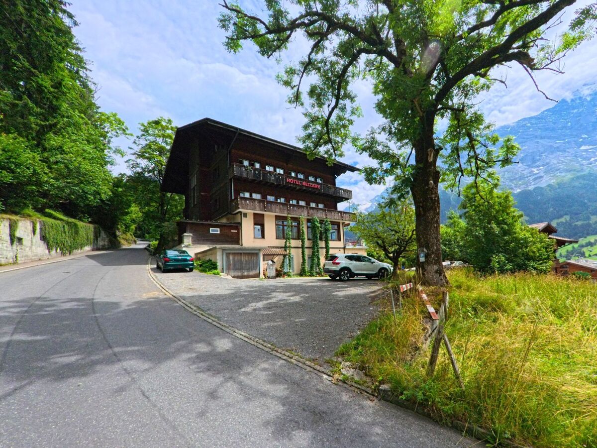 Am Waldrand von Grindelwald (3818, Kanton Bern) steht ein mehrstöckiges Hotel mit Holzfassade an einer ruhigen Strasse mit Blick auf die umliegende Berglandschaft.