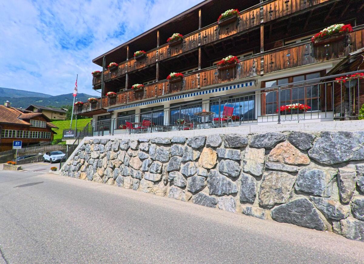 An einer leicht abschuessigen Strasse in Grindelwald (3818, Kanton Bern) befindet sich ein Hotel mit hölzernen Balkonen und einer Terrasse, die auf einer Stuetzmauer aus Naturstein liegt.