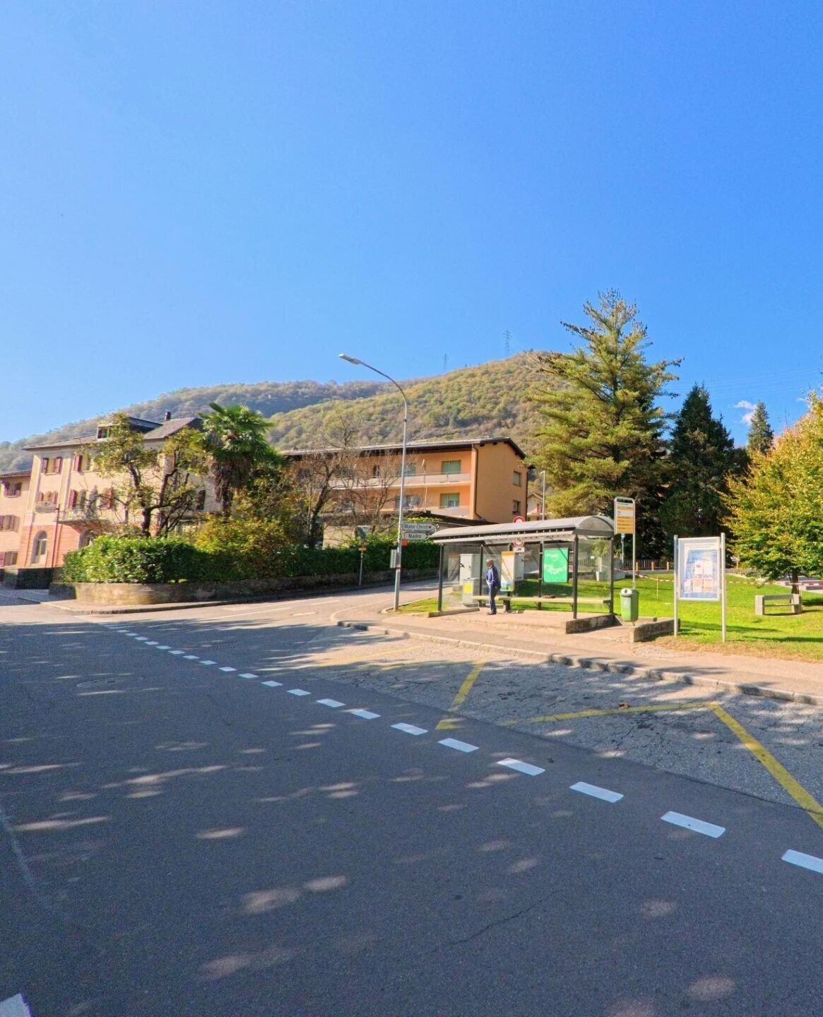 An einer Kreuzung mit Wartebereich zeigt sich in Grono (6537, Kanton Graubünden) eine Bushaltestelle neben Grünflächen und Wohnbauten vor dem bewaldeten Hang.