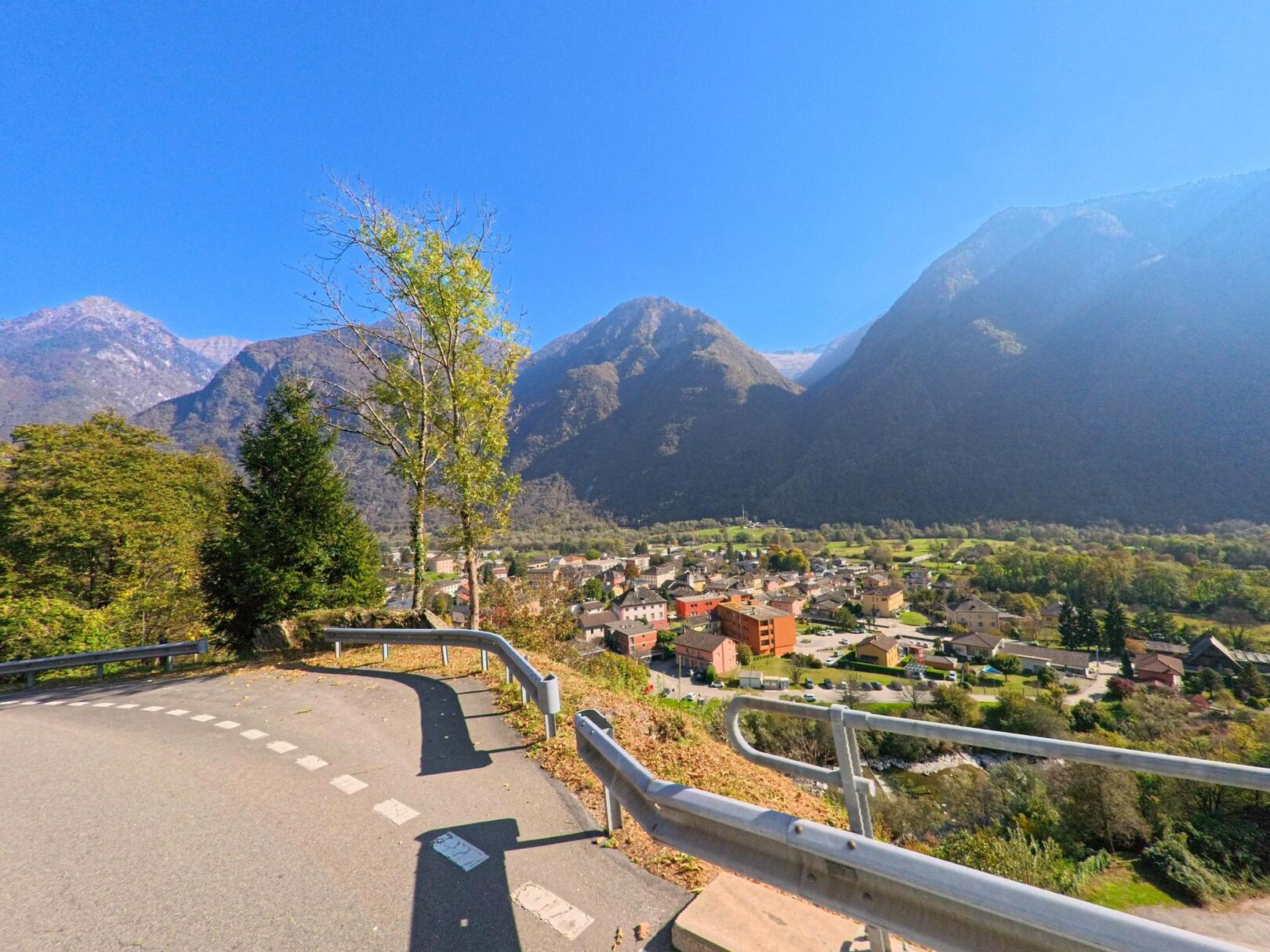 Von einer kurvigen Strasse oberhalb des Dorfes eröffnet sich der Blick auf Grono (6537, Kanton Graubünden) mit Wohnhäusern, Grünflächen und den steilen Berghängen im Hintergrund.
