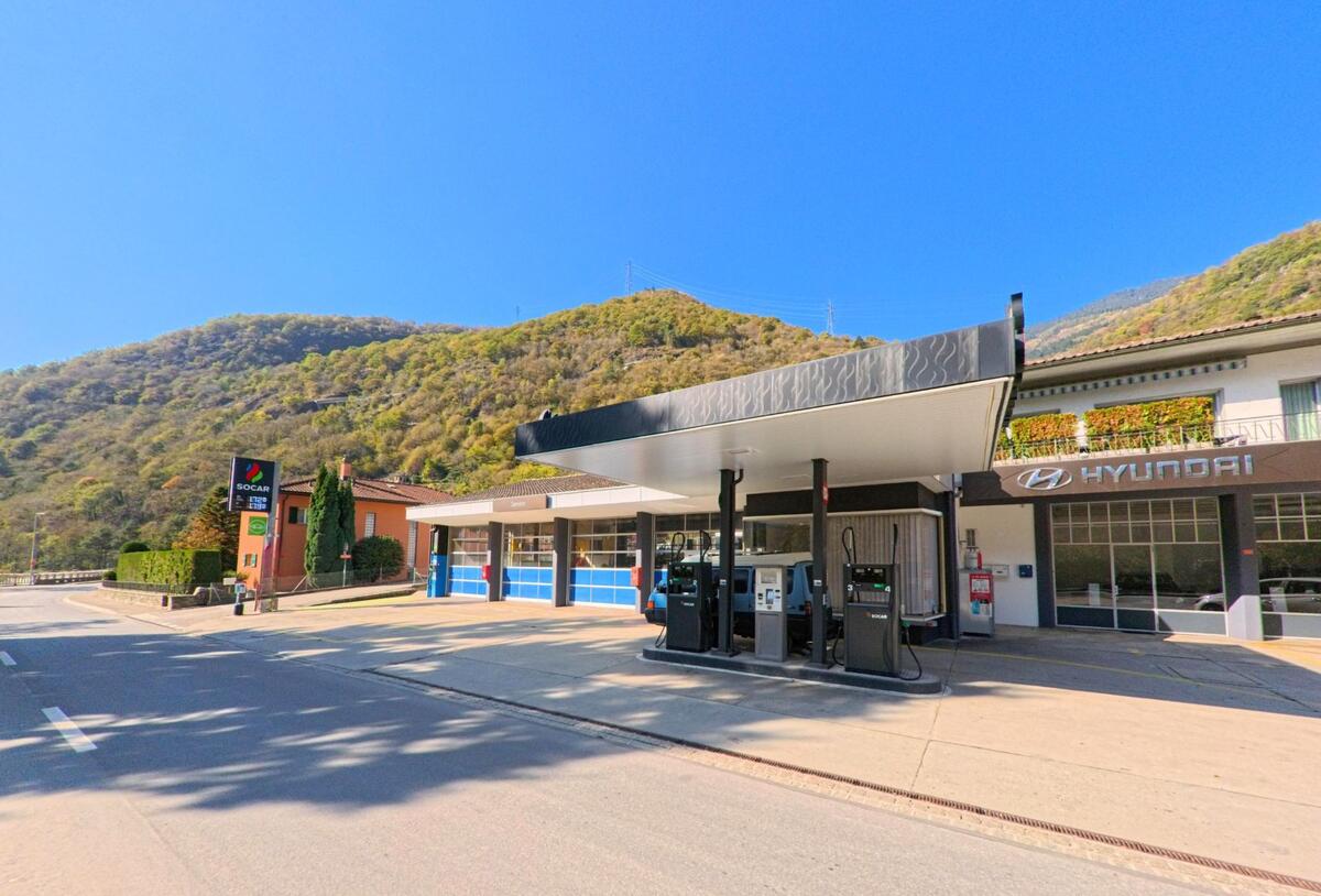 An der Hauptstrasse von Grono (6537, Kanton Graubünden) befindet sich eine Tankstelle mit Zapfsäulen neben Gewerbebauten vor bewaldeten Berghängen.