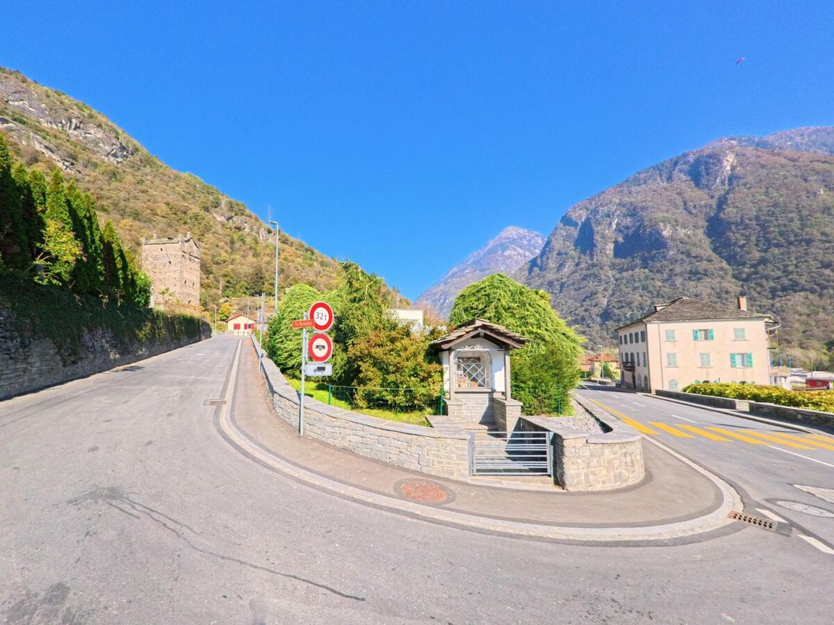 An einer Weggabelung in Grono (6537, Kanton Graubünden) steht eine kleine Wegkapelle zwischen Strasse, Begrünung und den steilen Berghängen des Tals.