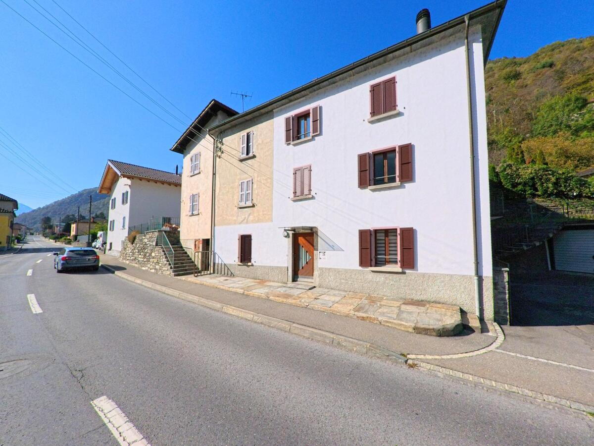 Entlang einer Dorfstrasse in Grono (6537, Kanton Graubünden) stehen traditionelle Wohnhäuser mit Fensterläden vor einem begrünten Hang.