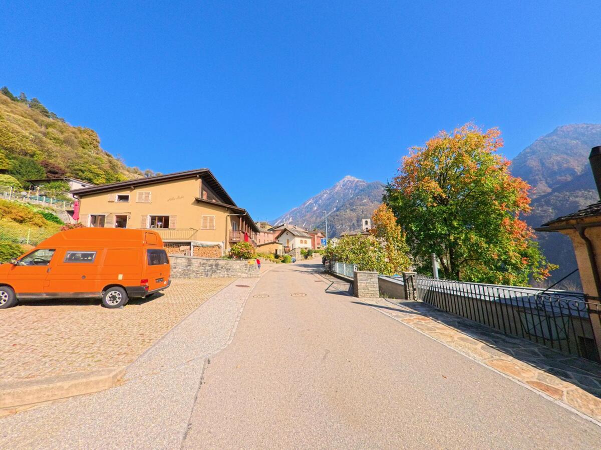Entlang einer leicht ansteigenden Quartierstrasse in Grono (6538, Kanton Graubünden) führen Wohnhäuser und herbstlich gefärbte Bäume den Blick in Richtung der umliegenden Berge.
