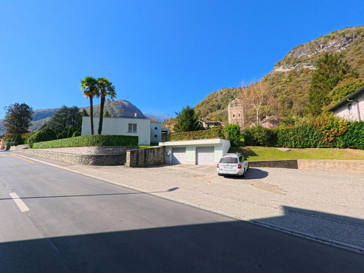An der Strasse in Grono (6556, Kanton Graubünden) steht ein modernes Wohnhaus mit Garten und Palmen, während im Hintergrund ein historischer Turm am bewaldeten Hang sichtbar ist.