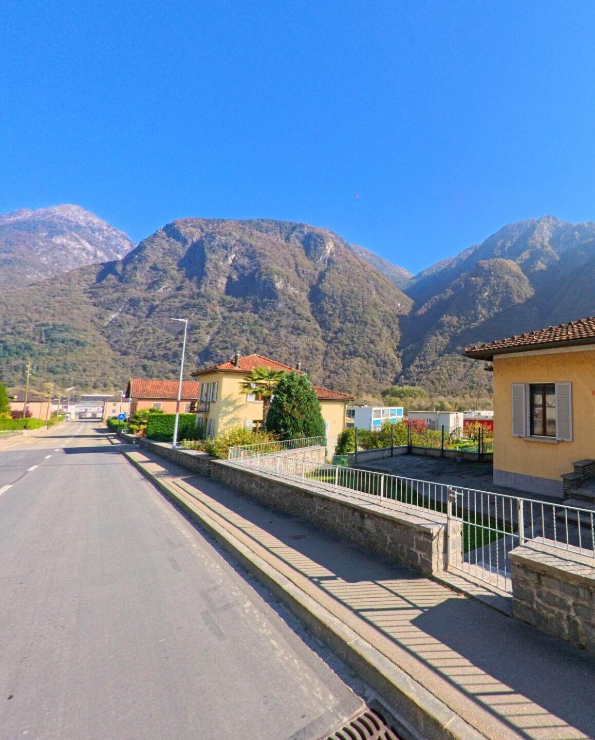 Entlang einer ruhigen Wohnstrasse in Grono (6556, Kanton Graubünden) liegen Einfamilienhäuser mit Gärten vor den steil ansteigenden Berghängen des Tals.