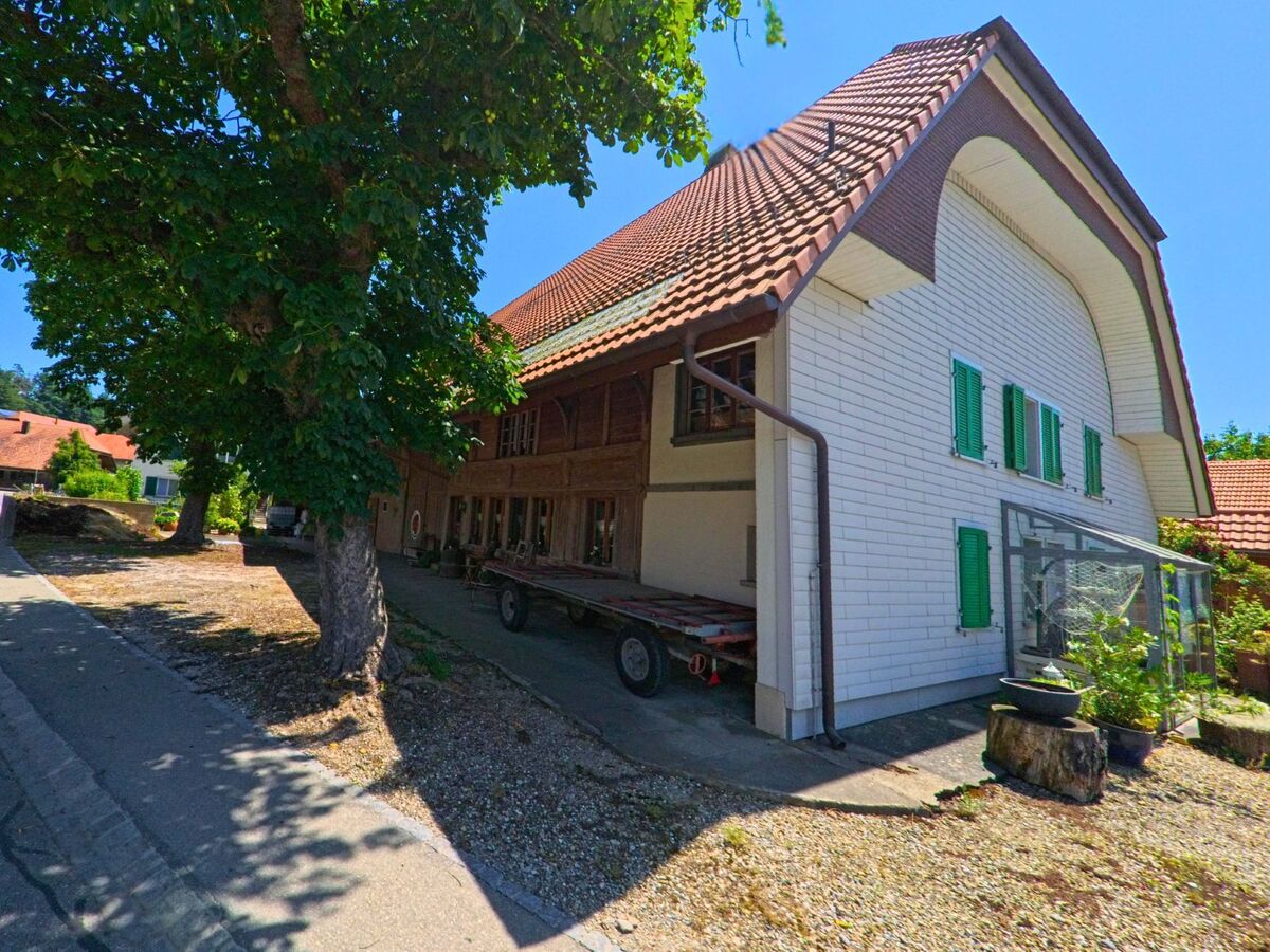 In Grossaffoltern (3257, Kanton Bern) steht ein traditionelles Bauernhaus mit grünen Fensterläden und grossem Vordach entlang eines schattigen Gehwegs.