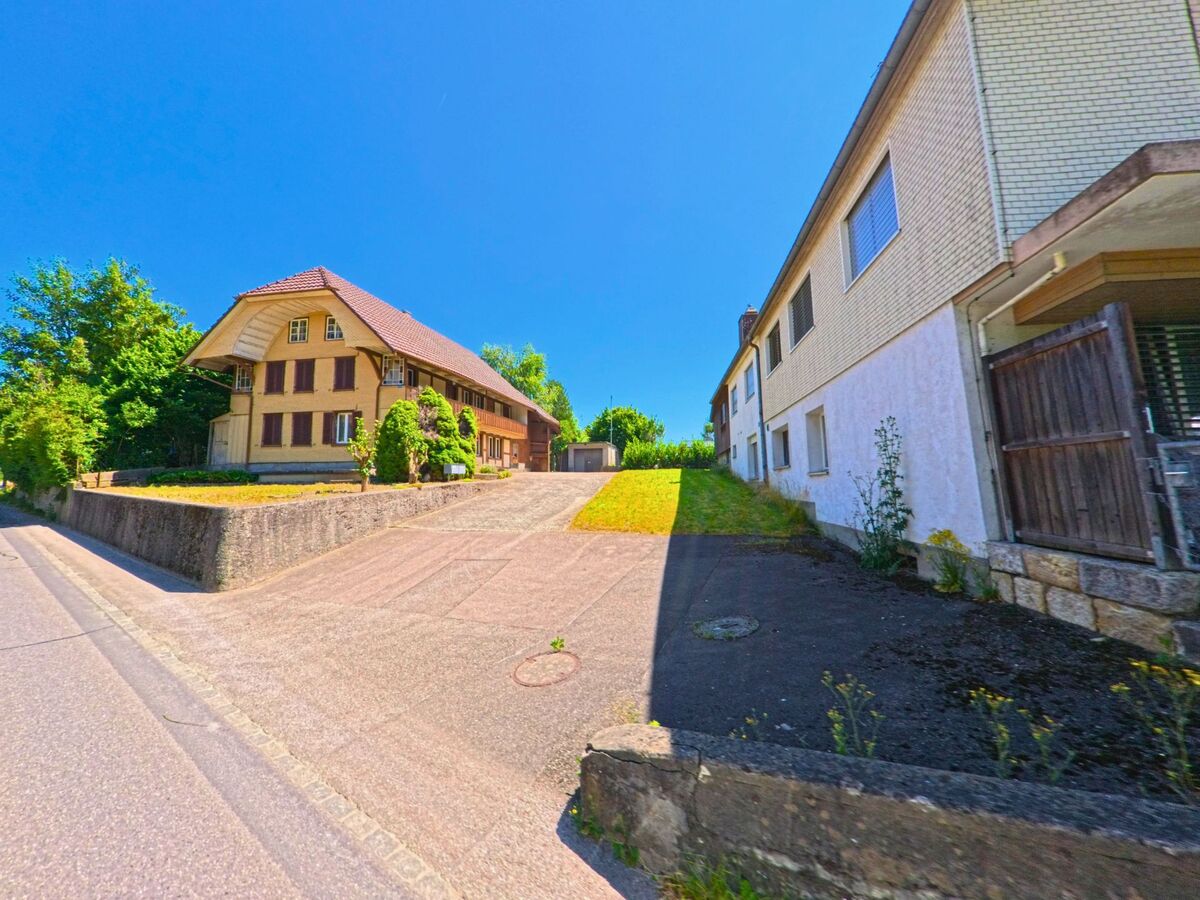 In Grossaffoltern (3257, Kanton Bern) führt eine Einfahrt zwischen einem alten Bauernhaus mit Walmdach und einem schlichten Mehrfamilienhaus leicht bergauf.